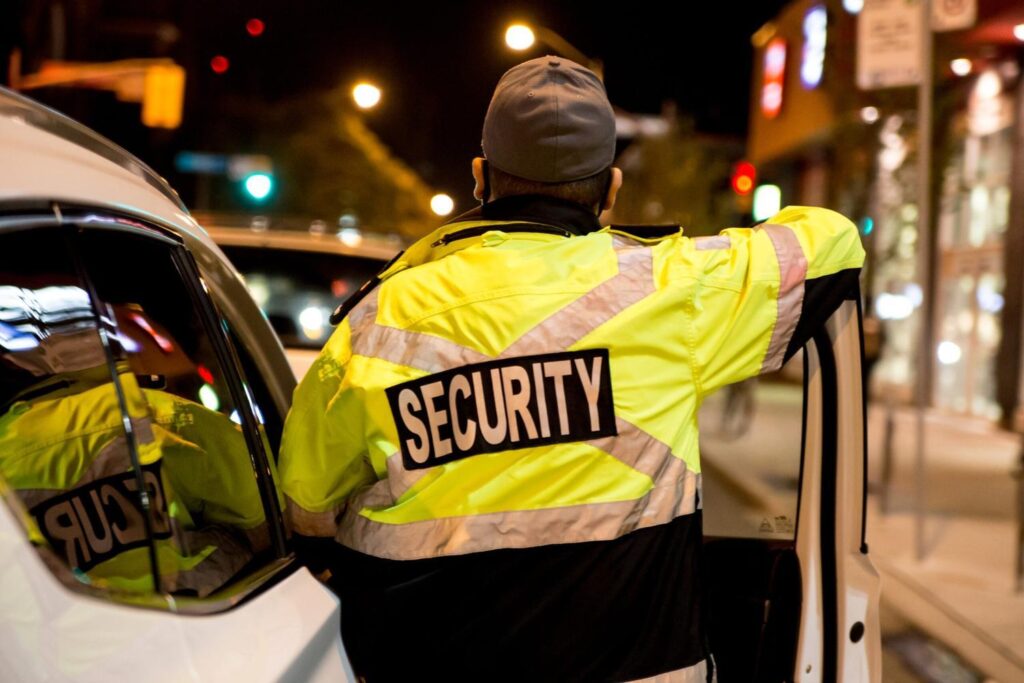 Mobile patrol security officer standing with a security vehicle outside a commercial building in the UK during an evening patrol.
