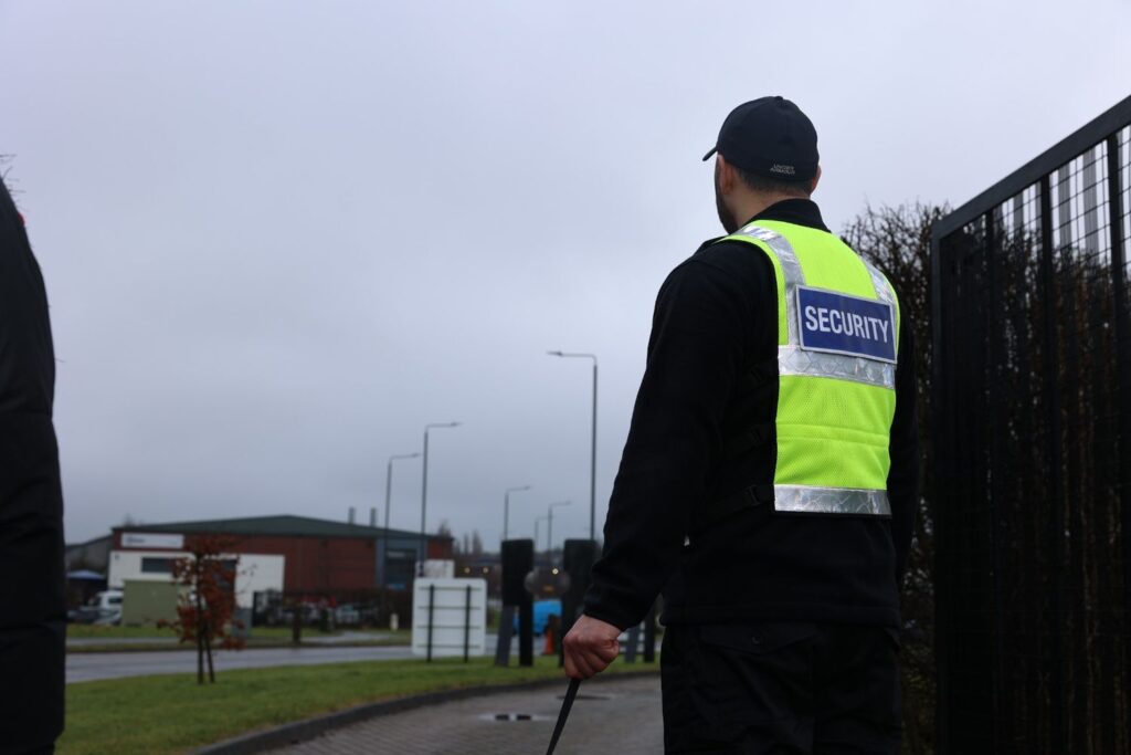 A professional security guard standing outside a building entrance during daytime in the UK, wearing a high-visibility uniform and observing the surroundings.